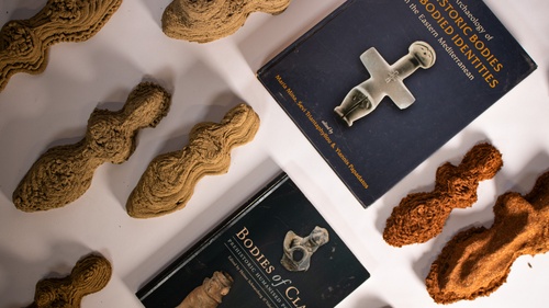 a photography of various human-like bodies made of printed earth-fiber, arranged with two reference books: bodies of clay and archeology of prehistoric bodies & embodied identities